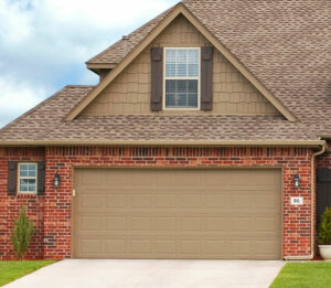 A two-story brick house with a gabled roof and tan shingles. The upper section has brown siding, and there are two brown shutters flanking an upper-story window. The tan garage door covers the bottom front, with a light above and a small trimmed plant on the side.