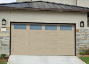 A beige garage door with horizontal panels and a row of rectangular windows at the top. The door is set in a stone and stucco wall. Two lantern-style lights are mounted on either side of the garage door. A driveway slopes down from the door.