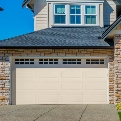 A beige-colored garage door with four small windows at the top is set in a house with stone and siding exterior. The house has a dark gray shingle roof and two upper windows. The driveway is clean and concrete, leading up to the garage.