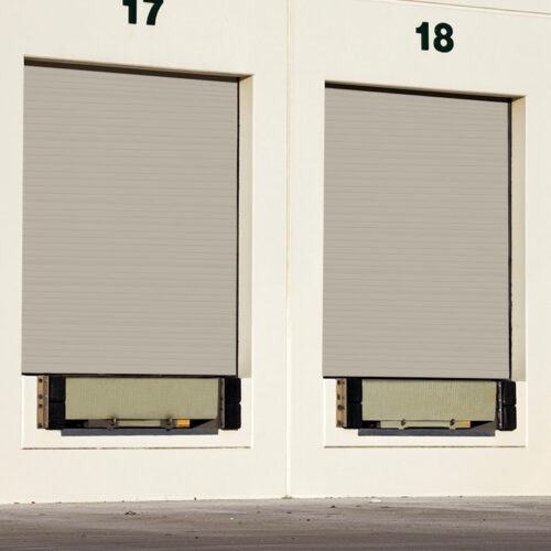 Two closed loading dock doors, numbered 17 and 18, are set in a white building. The doors are grey and roll-up style, with black bumpers at the base of each dock door. The concrete ground is visible in front of the doors.