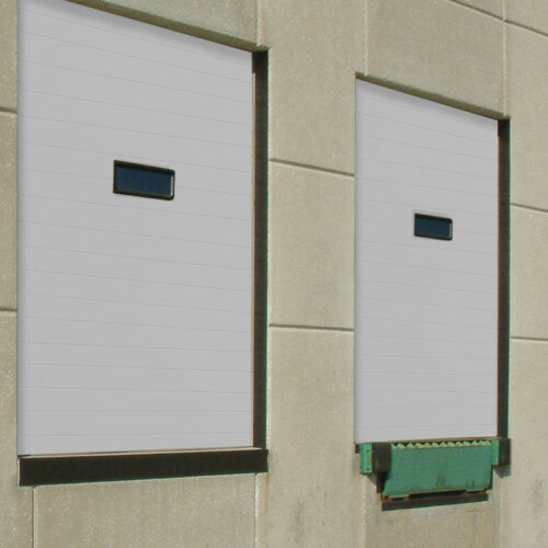 Two industrial loading dock doors on a beige concrete building wall. One door is closed, while the other is partially open, revealing a green dock leveler. Each door has a small rectangular window in the top center.