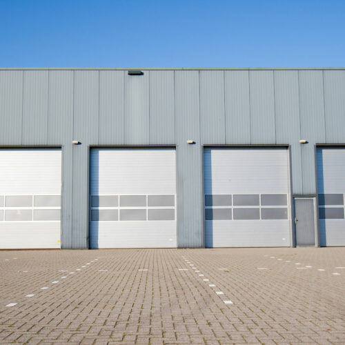 A large, gray industrial warehouse building with four closed roll-up doors sits under a clear blue sky. The foreground features a spacious, empty paved area with marked parking spaces and loading zones.