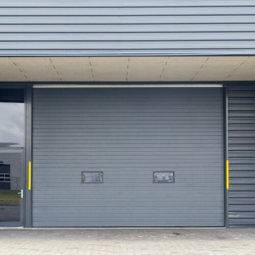 A large, gray industrial garage door with two small rectangular windows near the bottom. The surrounding building has a matching gray corrugated metal exterior. Yellow safety guards are mounted vertically on either side of the door. The ground is paved.