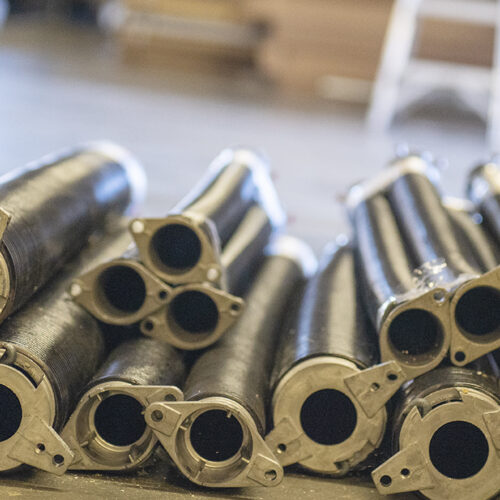 A pile of cylindrical metal pipes, stacked and arranged neatly on the ground in an industrial setting. Each pipe has a circular opening and mounting brackets on one end. The background is slightly blurred, indicating an indoor environment.