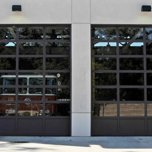 A modern fire station with two large glass panel garage doors. Inside, a red fire truck is partially visible. The exterior of the building is a combination of light-colored and textured concrete walls, with trees and other buildings reflected in the glass.