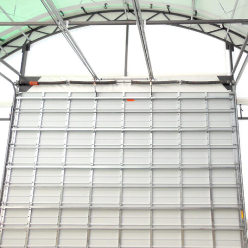 A large, industrial roll-up door in a warehouse or storage facility is partially open, revealing the metal tracks and the frame structure supporting it. The ceiling and walls of the facility are made of a translucent material, allowing light to filter through.