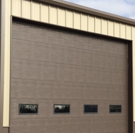 A large, closed industrial garage door in a beige and brown building. The door, in a dark brown color, has four small rectangular windows at the bottom. The building has a light beige upper section and a cloudy sky is visible above.