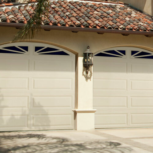 A house with a tiled roof features two adjacent white garage doors. Each door has a decorative window with a sunburst pattern at the top. An exterior light fixture is mounted on the wall between the garage doors. The driveway is plain and light-colored.