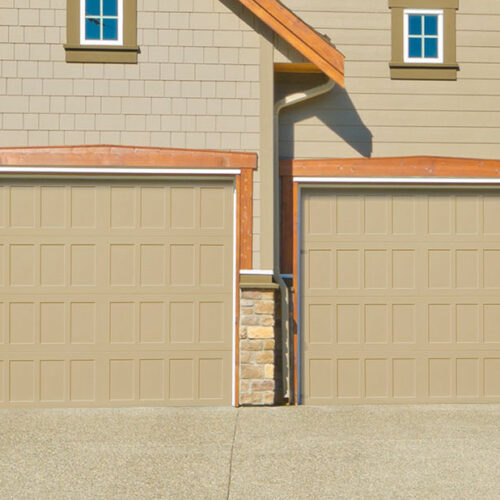 A tan house with two adjoining garages, each with light brown, recessed panel garage doors. The exterior features light brown siding, stone accents at the base, and wood trim around the garage doors and windows. The driveway is concrete.