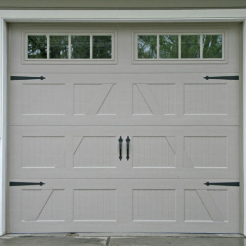 A beige garage door with black hinges and handles, featuring four small windows at the top. The door has a wood-paneled design and is set in a stone and siding wall. A lantern-style light fixture is mounted on the left side of the garage.