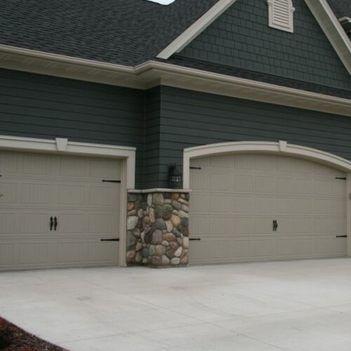 A residential home exterior with two beige, closed garage doors. The garage features stone accents on the lower portion of the walls and dark green horizontal siding on the upper portion. The driveway in front of the garage is made of smooth concrete.