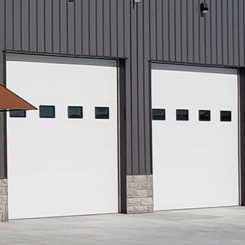 Two white garage doors in a dark gray building with small square windows aligned in a row on each door. The building features a combination of metal siding and a stone base at the bottom. A small awning extends over the left side door.