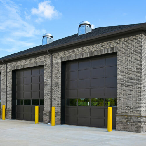 A modern warehouse building with two large, closed, dark brown garage doors. The building is made of light gray brick and has two ventilation ducts on the roof. Yellow bollards are positioned in front of each garage door, and the sky is clear and blue.