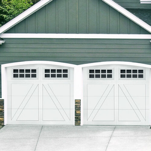 The image shows the front of a house with two adjacent white garage doors. Each door has a row of small square windows at the top and decorative horizontal and diagonal lines. The exterior wall is painted gray with white trim and stone accents at the bottom.