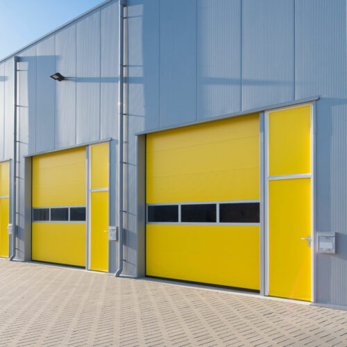 A modern industrial building featuring two large yellow roll-up doors with adjacent smaller pedestrian doors, set against a gray corrugated metal exterior. The ground is paved with light gray bricks, and a single light fixture is mounted on the wall.