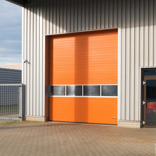 A large orange industrial garage door is set within a grey metal building. The door has a row of small, rectangular windows in the middle. The ground in front is paved, and part of a chain-link fence is visible on the left.
