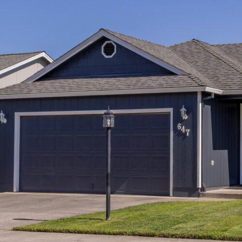 A suburban home with dark gray siding and a matching garage door. The house has a small front porch, two outdoor wall lanterns beside the garage, and the number 
