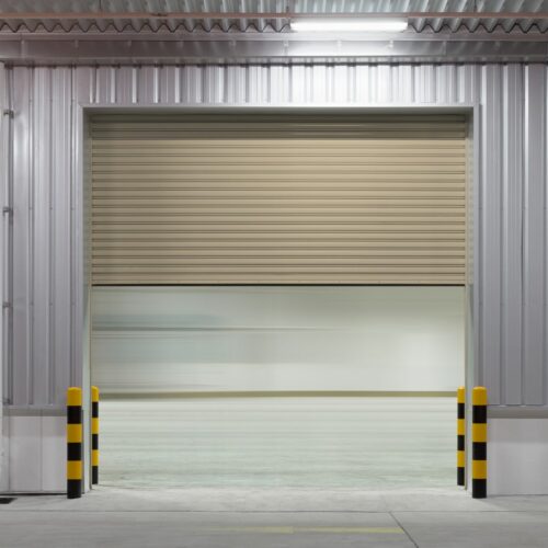 An open industrial garage door partially revealing a bright, spacious interior. The door is set within a corrugated metal wall, and two yellow and black striped bollards stand on either side of the entrance. The floor is clean and no objects are visible inside.