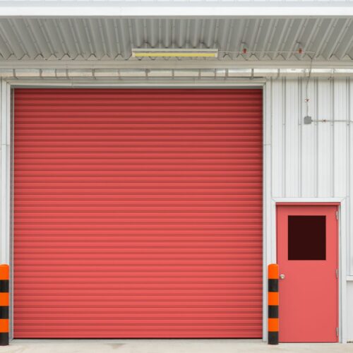 A large red rolling garage door next to a smaller red regular door on a white industrial building. There are black and orange safety bollards in front of the doors, and the roof has a corrugated metal structure.