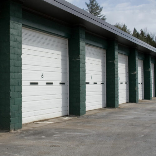 A row of storage unit garages with green cinder block walls and white roll-up doors, each numbered sequentially from 6 to 9. The garages are situated in an outdoor location, with trees visible in the background and a wet concrete floor in the foreground.