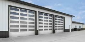 A modern commercial building with four large, closed garage doors. The structure is white with dark trim and has a flat roof. The surrounding area is paved with concrete, and the sky above is clear and blue.