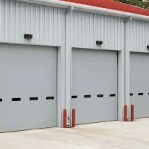 A row of three closed gray garage doors in a metal building, each with black panels and small windows. The building has a light fixture above each door and is set on a concrete surface with red protective bollards in front of each door.