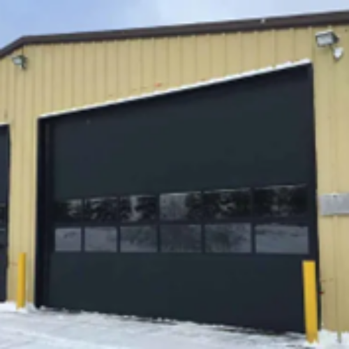 A beige industrial building with two large black garage doors and yellow posts in front. The ground is covered with snow, and the sky is overcast.