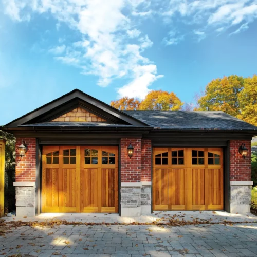 A double garage with wooden doors and a brick and stone exterior is pictured on a sunny day. The driveway is paved with grey stones and there are autumn trees in the background with orange and yellow leaves. The sky is blue with scattered clouds.