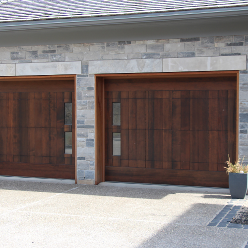 A modern residential garage with two wooden doors set into a stone facade. The driveway is paved with light-colored concrete, and there is a small planter with a tall, thin plant next to the right garage door.