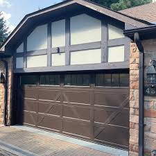 A brown garage door with a crossbuck design is set into a brick exterior. The upper portion of the garage door has windows, and above it is a facade with a Tudor-style pattern in dark brown and off-white. A lantern-style light fixture is attached to the brick wall.